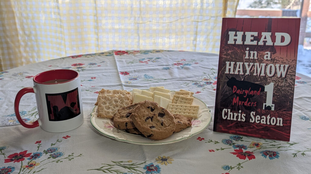 Welcome image of tablecloth with coffee, plate of cookies, and Book 1 of the Dairyland Murders Series